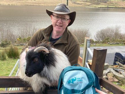 Ein lächelnder Mann mit Brille trägt einen Hut und hält ein Schaf auf einem Zaun, während eine malerische Landschaft im Hintergrund zu sehen ist.