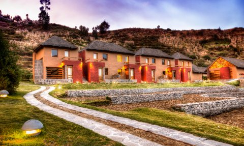 Die malerische Lodge mit strohgedeckten Dächern liegt in einer sanften Hügellandschaft, umgeben von üppigem Grün.