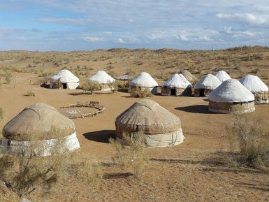 Eine Gruppe traditioneller Jurten steht in einer trockenen, sandigen Landschaft unter einem klaren Himmel.