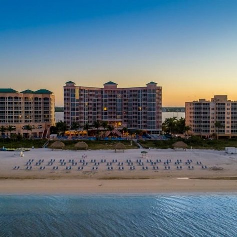 Ein Blick auf den Fort Myers Beach mit mehreren Gebäuden und Liegen am Strand, während die Sonne untergeht.