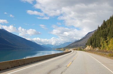 Eine kurvenreiche Straße entlang eines ruhigen Sees, umgeben von majestätischen Bergen und bewaldeten Hängen.