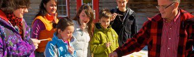 Eine Gruppe von Kindern und Erwachsenen genießt fröhlich eine süße Leckerei im Freien, während die Sonne scheint.