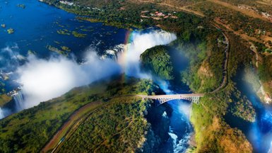 Ein atemberaubender Blick auf die Victoriafälle, umgeben von üppigem Grün und einem strahlenden Regenbogen über dem Wasserfall.