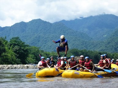 Eine Gruppe von Menschen in gelben Rafts, während ein Guide fröhlich in der Luft springt, umgeben von grünen Bergen.