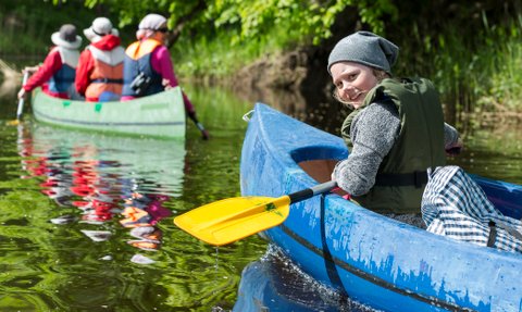 Ein fröhliches Kind paddelt in einem blauen Kajak, während andere Kajakfahrer im Hintergrund auf einem ruhigen Gewässer fahren.
