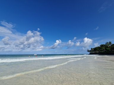 Ein klarer Strand mit sanften Wellen, unter einem strahlend blauen Himmel, der von weißen Wolken durchzogen ist.