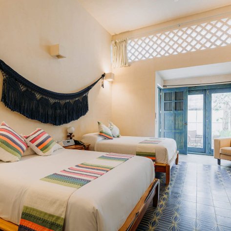 Ein helles Zimmer mit zwei Betten, bunten Kissen und einer makramee Hängematte an der Wand, strahlt Gemütlichkeit aus.