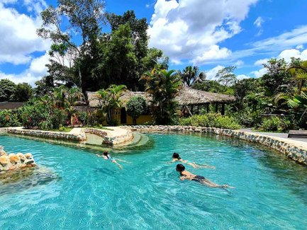 Drei Kinder schwimmen fröhlich in einem klaren Pool, umgeben von üppigem Grün und strahlend blauem Himmel.