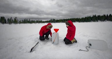 Zwei Personen in roten Jacken stehen auf einer verschneiten Fläche und bereiten sich auf das Eisangeln vor.