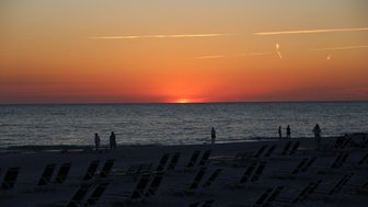 Ein atemberaubender Sonnenuntergang über dem Meer, während Menschen am Strand spazieren und Liegen im Vordergrund stehen.