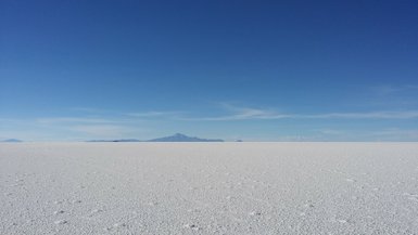 Eine weite, weiße Salzfläche erstreckt sich bis zum Horizont, unter einem strahlend blauen Himmel mit wenigen Wolken.