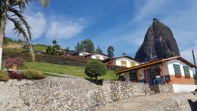 Bunte Häuser mit blühenden Pflanzen vor dem beeindruckenden Felsen El Peñol unter einem strahlend blauen Himmel.