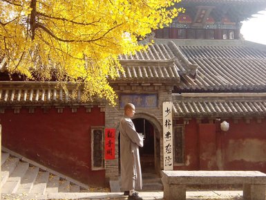 Ein Mönch in traditioneller Kleidung steht nachdenklich vor einem alten Kloster, umgeben von leuchtend gelben Blättern.