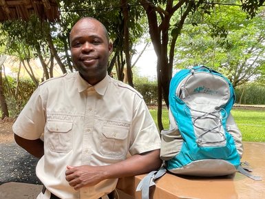 Ein lächelnder Mann in khakifarbener Kleidung steht neben einem Rucksack auf einem Tisch aus Stein in einer grünen Umgebung.