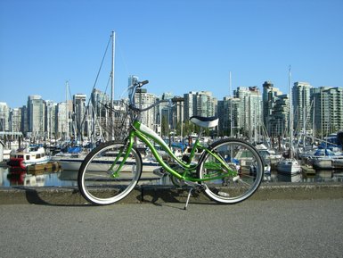 Ein grünes Fahrrad steht auf dem Gehweg, mit einer Marina und modernen Hochhäusern im Hintergrund.