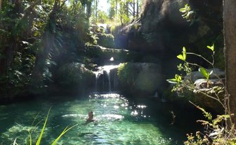 Ein klarer Naturpool umgeben von üppigem Grün und sanften Wasserfällen, in dem eine Person entspannt schwimmt.