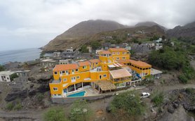 Blick auf ein leuchtend orangefarbenes Gebäude mit Pool, umgeben von sanften Hügeln und dem Meer im Hintergrund.