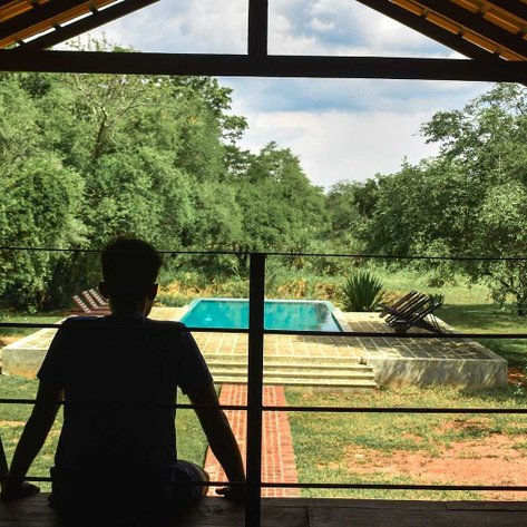 Ein junger Mann sitzt auf einer Veranda und blickt auf einen ruhigen Pool, umgeben von üppigem Grün und Bäumen.