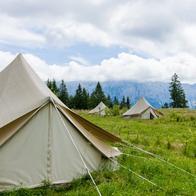 Eine Gruppe von Zelten steht auf einer grünen Wiese, umgeben von hohen Bäumen und majestätischen Bergen im Hintergrund.