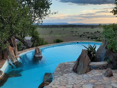 Ein glitzernder Pool umgeben von Felsen und Pflanzen, mit einer weiten Aussicht auf die Savanne und den Himmel bei Sonnenuntergang.