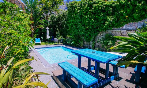 Ein einladender Poolbereich mit einem blauen Tisch und Bänken, umgeben von üppigem Grün und einem sonnigen Himmel.