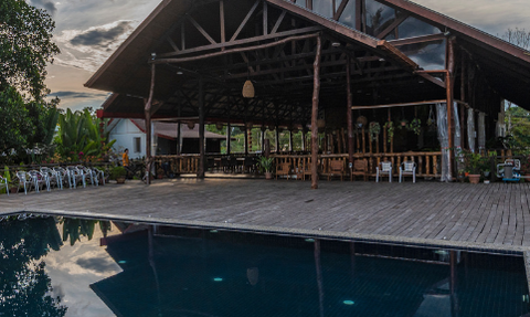 Eine rustikale Lodge mit einer großen Veranda steht über einem spiegelglatten Pool, der die Wolken reflektiert.