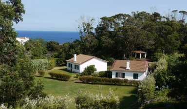 Zwei weiße Häuser mit braunen Dächern stehen in einem grünen Garten, umgeben von Bäumen und Blick auf das blaue Meer.