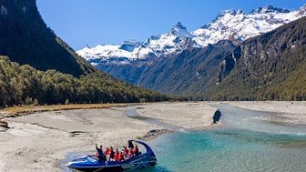 Ein Jetboot mit mehreren Passagieren gleitet über den klaren Fluss, umgeben von majestätischen Bergen und strahlend blauem Himmel.
