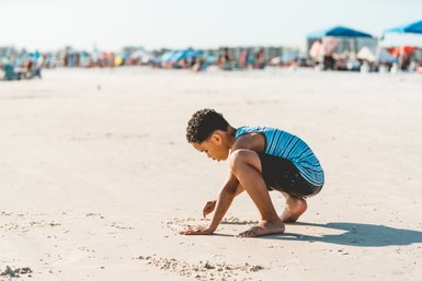 Ein Junge mit lockigem Haar sitzt am Strand und gräbt mit seinen Händen im Sand, während die Sonne scheint.