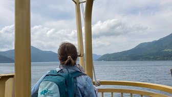 Eine Person mit einem Rucksack steht an einem Geländer und blickt auf einen ruhigen See, umgeben von Bergen.