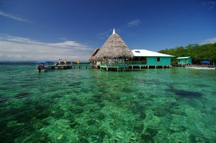 Ein malerisches Wasserhaus mit Strohdach steht auf einem Holzsteg über dem klaren, türkisfarbenen Wasser.