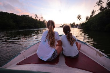 Zwei Mädchen sitzen auf einem Boot und blicken auf den Sonnenuntergang über dem Wasser, umgeben von Palmen.