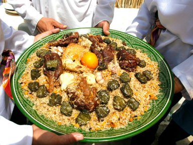 Ein großer, grüner Teller mit duftendem Plov, garniert mit Hähnchen, Lamm und gefüllten Weinblättern, wird von mehreren Händen gehalten.