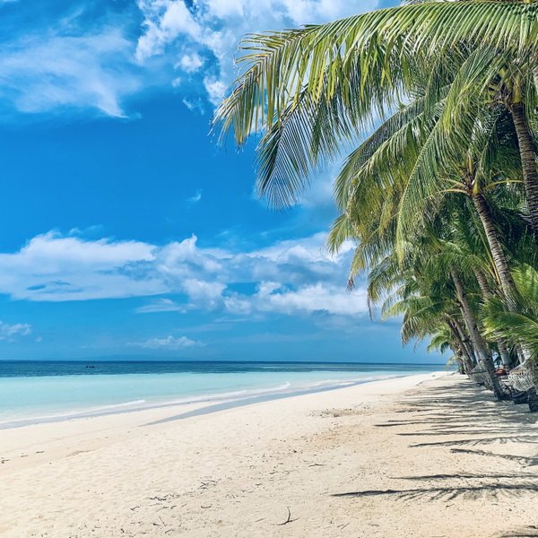 Ein ruhiger Strand mit feinem, weißen Sand erstreckt sich unter einem strahlend blauen Himmel und sanften Wolken.