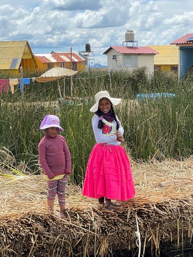 Zwei Mädchen stehen auf einem Schilfweg, umgeben von grünen Pflanzen und bunten Häusern am Titicaca-See.