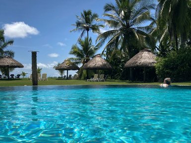 Ein klarer, blauer Swimmingpool spiegelt den strahlenden Himmel wider, umgeben von schattenspendenden Palmen und Hütten.