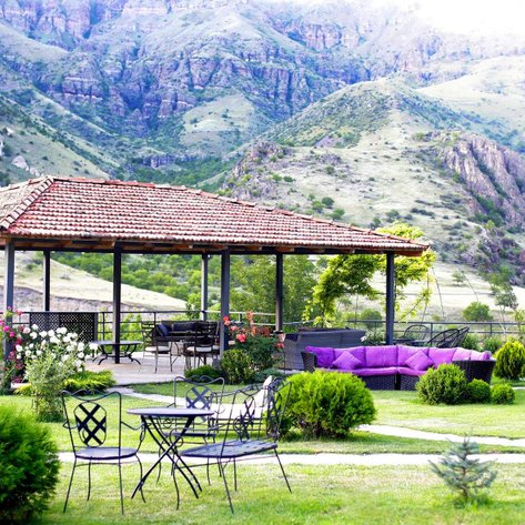 Ein einladender Pavillon mit einem schattigen Dach steht in einer grünen Landschaft, umgeben von sanften Hügeln und Bergen.