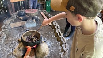 Ein Junge rührt mit einem Holzlöffel in einem Topf über einem offenen Feuer, während er konzentriert zusieht.