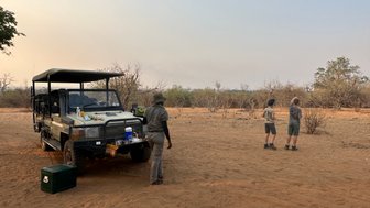 Ein Geländewagen steht auf sandigem Boden, während zwei Kinder in der Ferne die Umgebung beobachten.