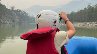 Ein Kind in einem roten Schwimmwesten und Helm paddelt auf einem ruhigen Fluss, umgeben von sanften Hügeln und Nebel.