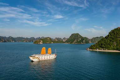 Ein traditionelles Boot mit orangefarbenen Segeln gleitet ruhig durch das klare Wasser der Halong-Bucht, umgeben von grünen Inseln.