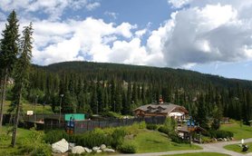 Eine malerische Lodge umgeben von hohen Bäumen und sanften Hügeln unter einem strahlend blauen Himmel mit Wolken.