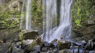 Ein Mensch steht auf einem großen Felsen und betrachtet einen majestätischen Wasserfall, umgeben von üppigem Grün.