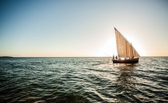 Ein traditionelles Dhow-Boot segelt majestätisch über das ruhige Wasser, während die Sonne am Horizont untergeht.