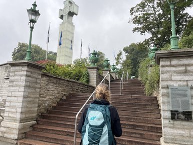 Eine Person mit einem Rucksack geht eine steinerne Treppe hinauf, umgeben von grünen Laternen und Flaggen.
