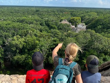 Drei Personen sitzen auf einer Mauer und blicken auf eine grüne Landschaft mit alten Ruinen im Hintergrund.