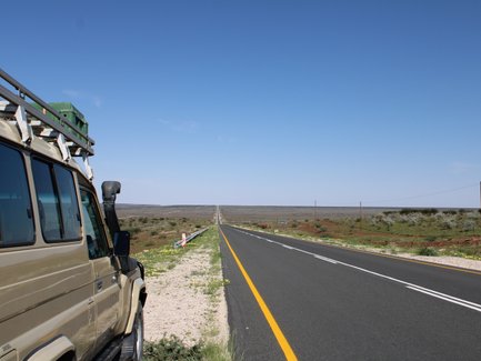 Ein Geländewagen steht an einer endlosen Straße in Namibia, umgeben von karger Landschaft und klarem Himmel.
