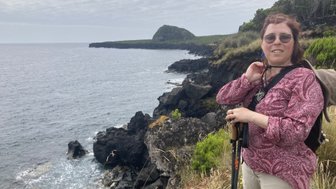 Eine Frau mit Brille steht an einer Küste, umgeben von Felsen und dem Meer, während sie einen Wanderstock hält.
