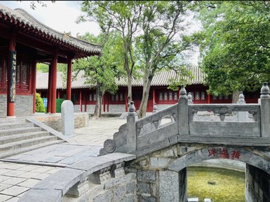 Eine steinerne Brücke führt über einen klaren Teich, umgeben von üppigem Grün und traditioneller Architektur.