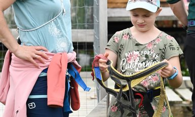 Ein Mädchen hält einen Klettergurt in der Hand und schaut konzentriert darauf, während eine Frau neben ihr steht.